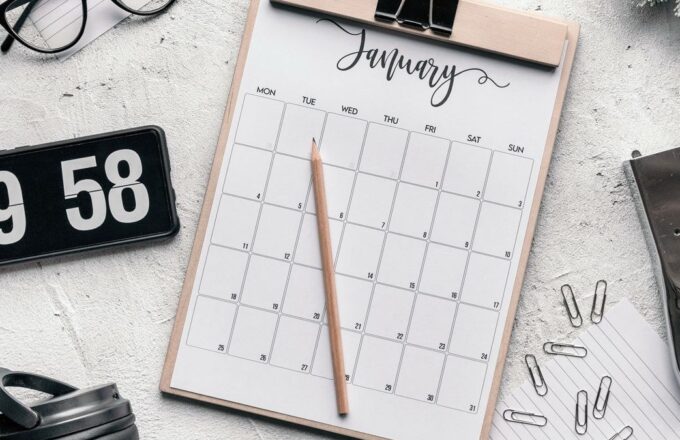 Monatskalender auf einem Klemmbrett mit Bleistift, daneben Brille, Uhr und Büroklammern