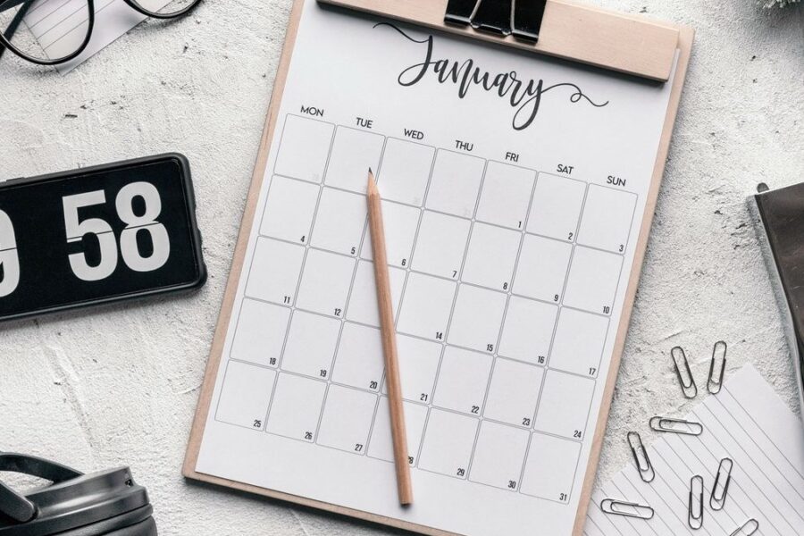 Monatskalender auf einem Klemmbrett mit Bleistift, daneben Brille, Uhr und Büroklammern