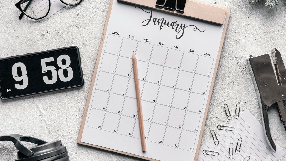 Monatskalender auf einem Klemmbrett mit Bleistift, daneben Brille, Uhr und Büroklammern
