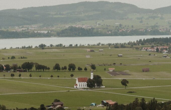 Weiße Wallfahrtskirche zwischen Wiesen im Voralpenland, dahinter ein See und bewaldete Hügel