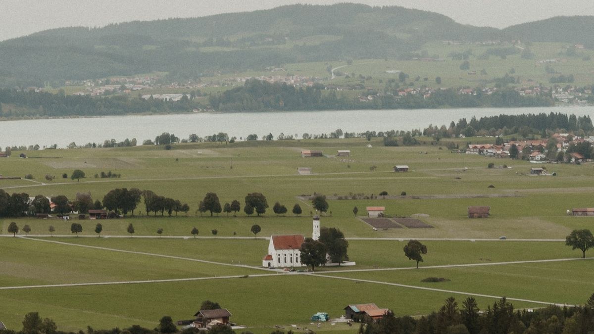Weiße Wallfahrtskirche zwischen Wiesen im Voralpenland, dahinter ein See und bewaldete Hügel