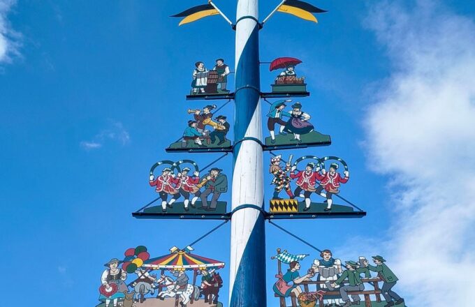Weiß-blauer Maibaum in München mit bemalten Figuren aus Brauchtum, Musik und Festen vor blauem Himmel