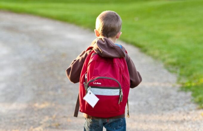 Kind mit rotem Schulrucksack läuft auf einem Kiesweg neben einer Wiese