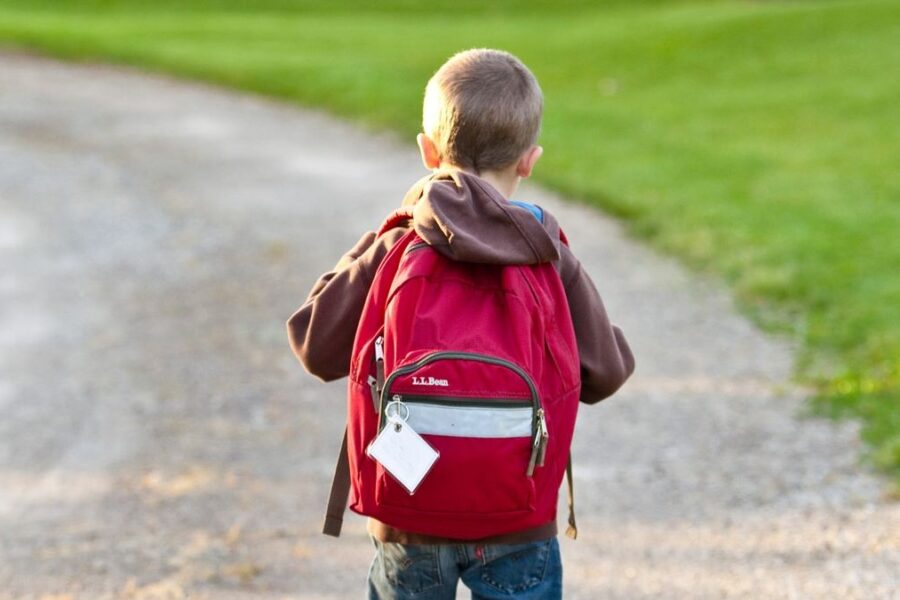 Kind mit rotem Schulrucksack läuft auf einem Kiesweg neben einer Wiese