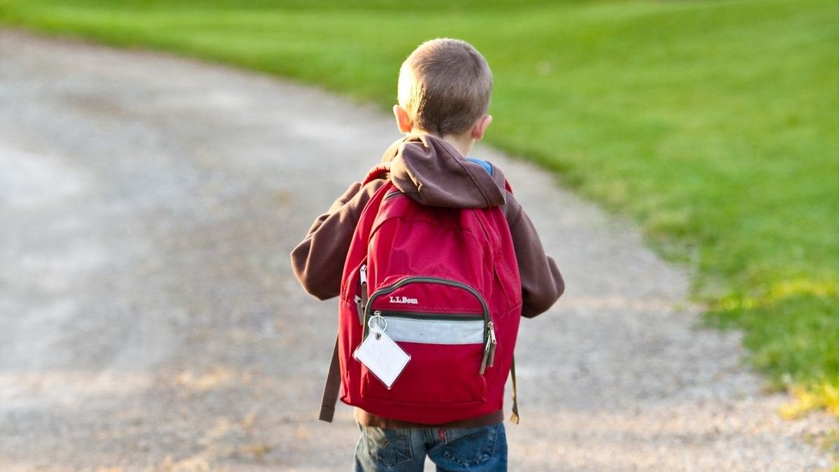 Kind mit rotem Schulrucksack läuft auf einem Kiesweg neben einer Wiese