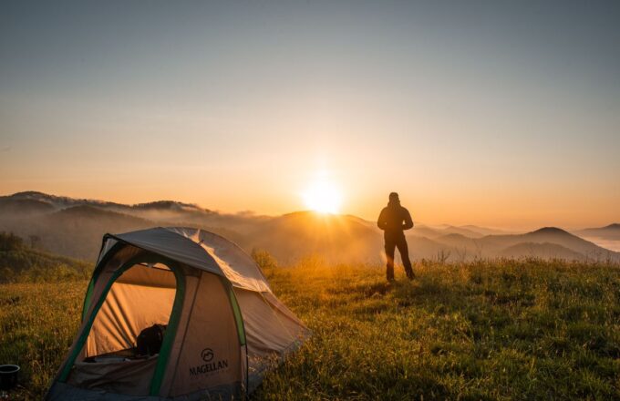Zelt auf einer Bergwiese bei Sonnenaufgang, daneben eine Person mit Blick über nebelige Hügel