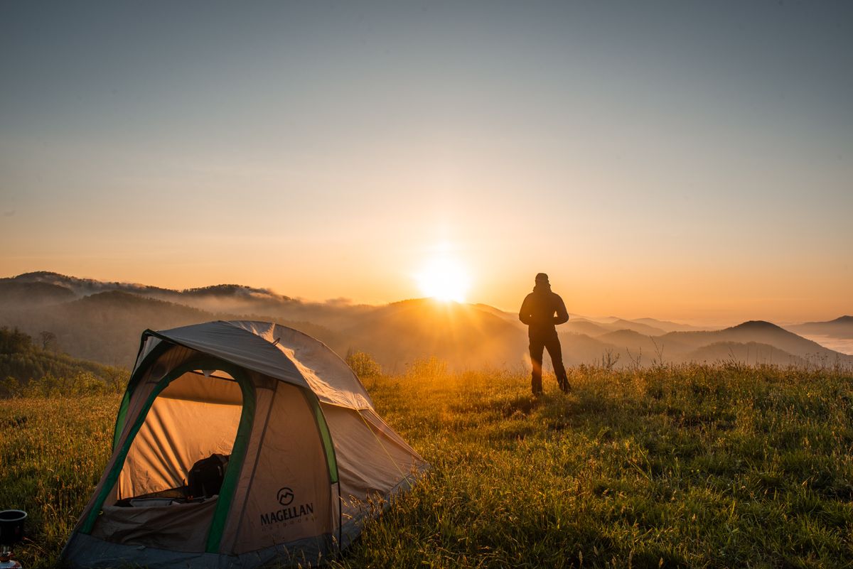 Zelt auf einer Bergwiese bei Sonnenaufgang, daneben eine Person mit Blick über nebelige Hügel